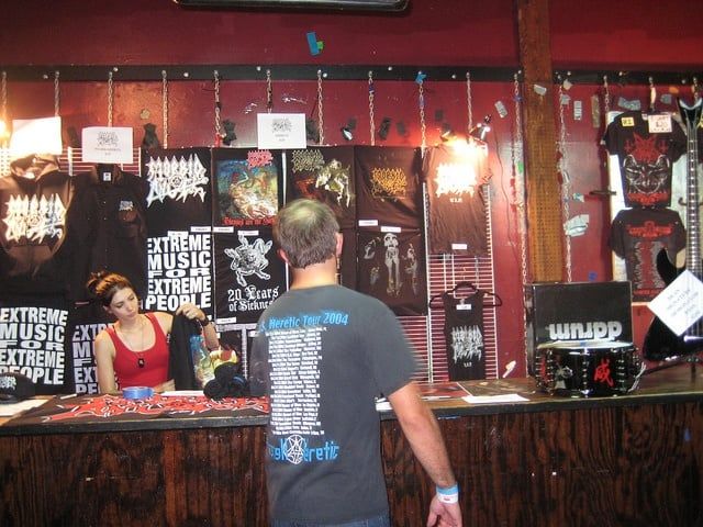 Merchandise-Stand einer Metal-Band mit T-Shirts und Postern bei einem Konzert.